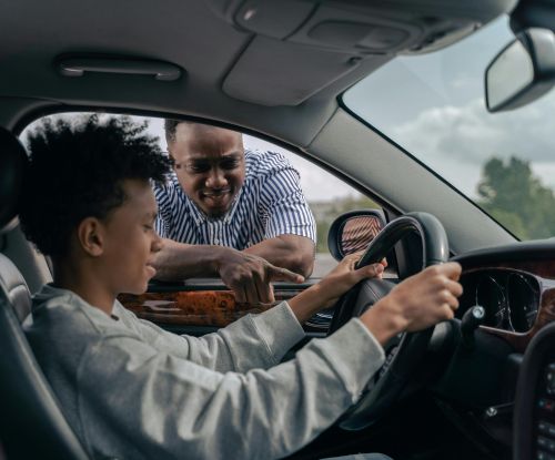 Young boy sitting at the wheel while father instructs from outside, learning to drive.